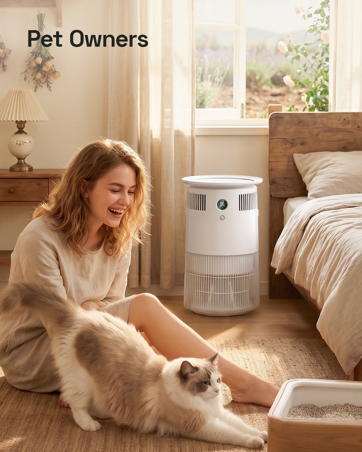 A woman sits on a bedroom floor smiling next to a fluffy cat, with a white pet Airdog air cleaner standing beside her. The scene is warmly lit by a window, with a bed, lamp, and cat litter box visible.