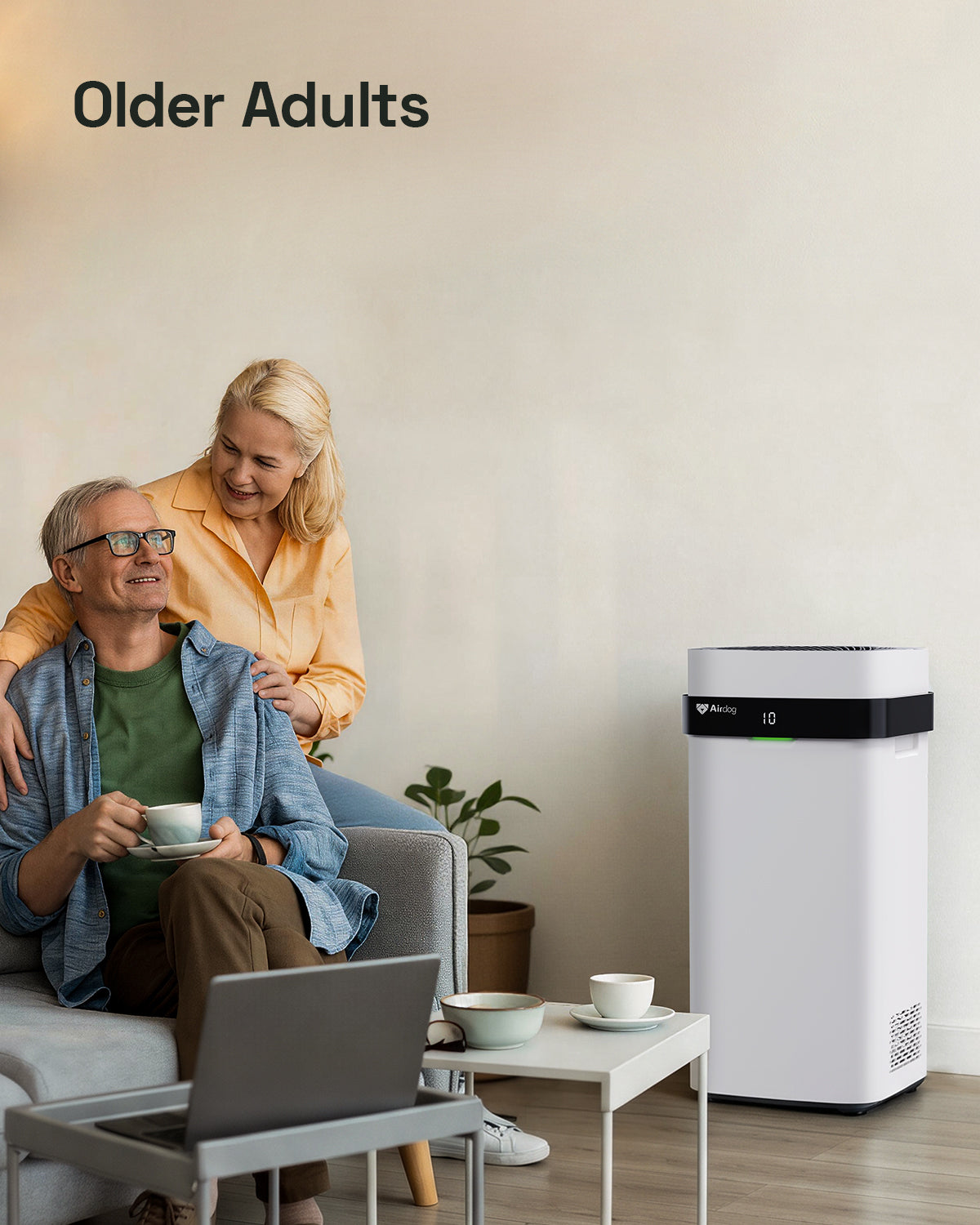 A white and black Airddog air cleaner stands on a light wood floor in a bright living room, next to a small table with cups and a laptop. Two older adults are seated on a sofa in the background, with the text 'Older Adults' in the top left corner.