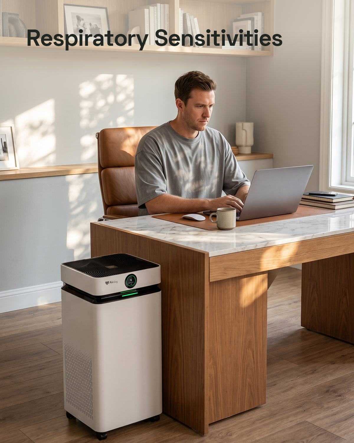 A man sits at a wooden desk using a laptop in a bright home office, with a white and black Airdog air purifier placed on the floor nearby. The text 'Respiratory Sensitivities' appears at the top of the image.