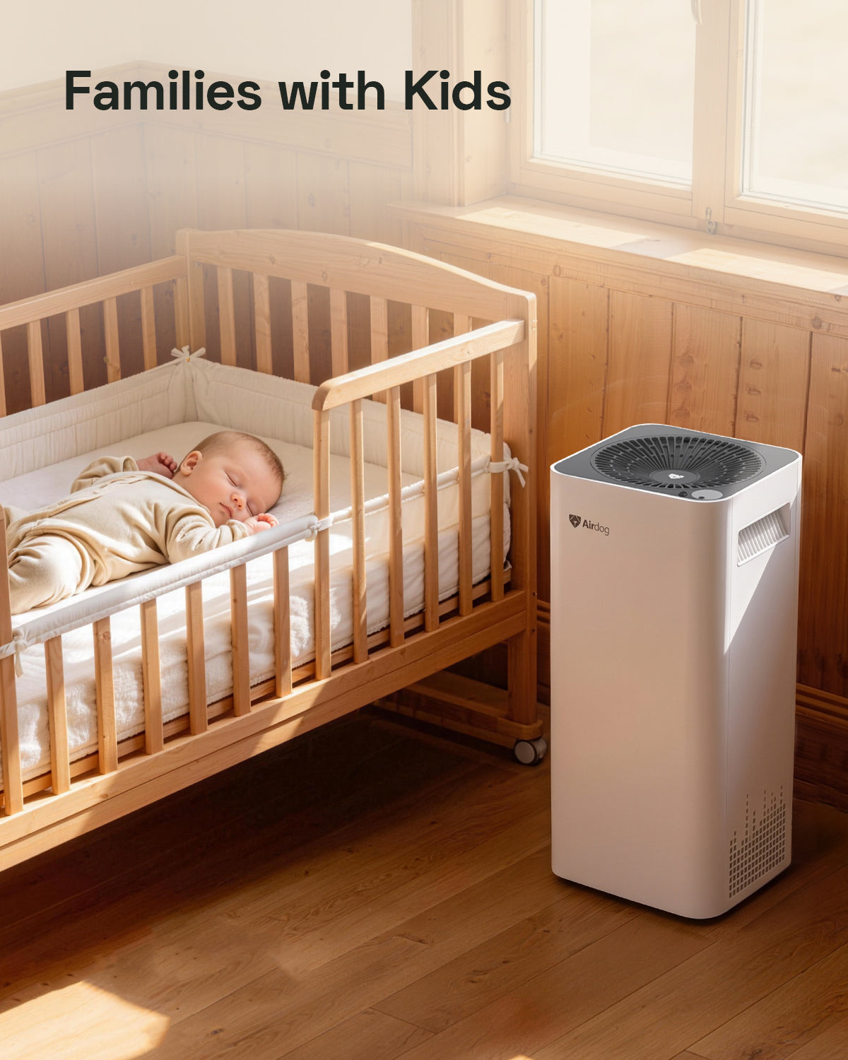 A white and gray Airdog air cleaner stands on a wooden floor next to a wooden baby crib with a sleeping baby inside, under warm window light. The text 'Families with Kids' is displayed at the top left.